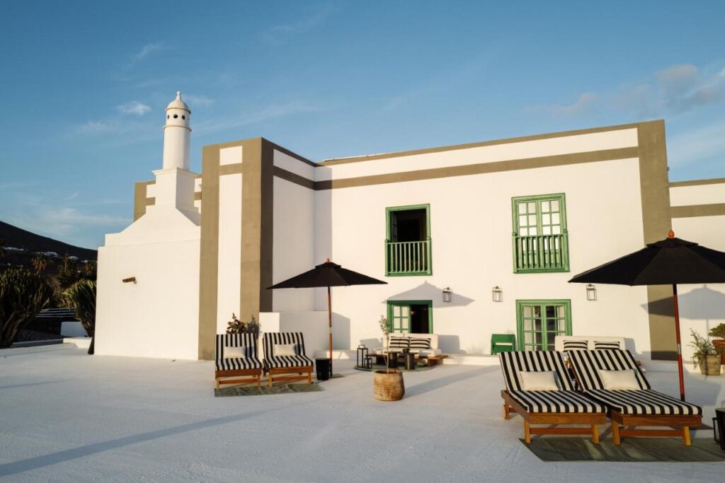 Unique Adults Only Hotels in Lanzarote 3 A modern white building with green-trimmed windows and doors, this adults-only hotel in Lanzarote features striped lounge chairs, black umbrellas, and outdoor seating on a spacious sunny terrace.