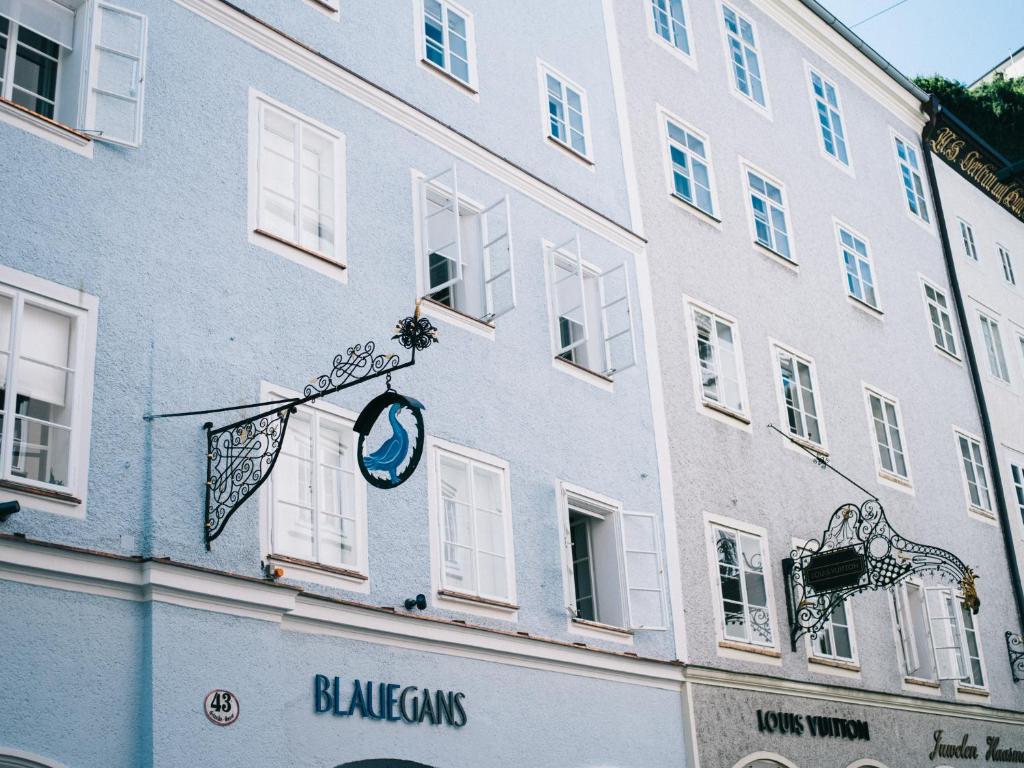 Unique Boutique Hotels in Salzburg 35 The image shows pale blue and white buildings with decorative wrought iron signs. Open windows line the walls, and shop signs read BLAUEGANS and LOUIS VUITTON—evoking the charm of boutique hotels in Salzburg. The scene is bright and airy.