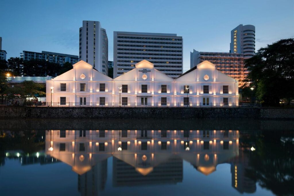 13 Most Unique Hotels in Singapore for an Incredible Holiday 16 A white, colonial-style building with illuminated windows, one of the unique hotels in Singapore, is reflected in a calm river at dusk, with modern high-rise buildings and trees visible in the background.