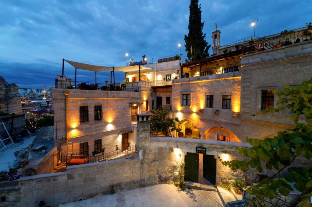 Most Authentic Cave Hotels in Cappadocia 2 A historic stone building with warm yellow lights glows at dusk. Reminiscent of cave hotels in Cappadocia, the multi-level structure has terraces, arched windows, greenery, and overlooks a scenic view under a cloudy blue sky.