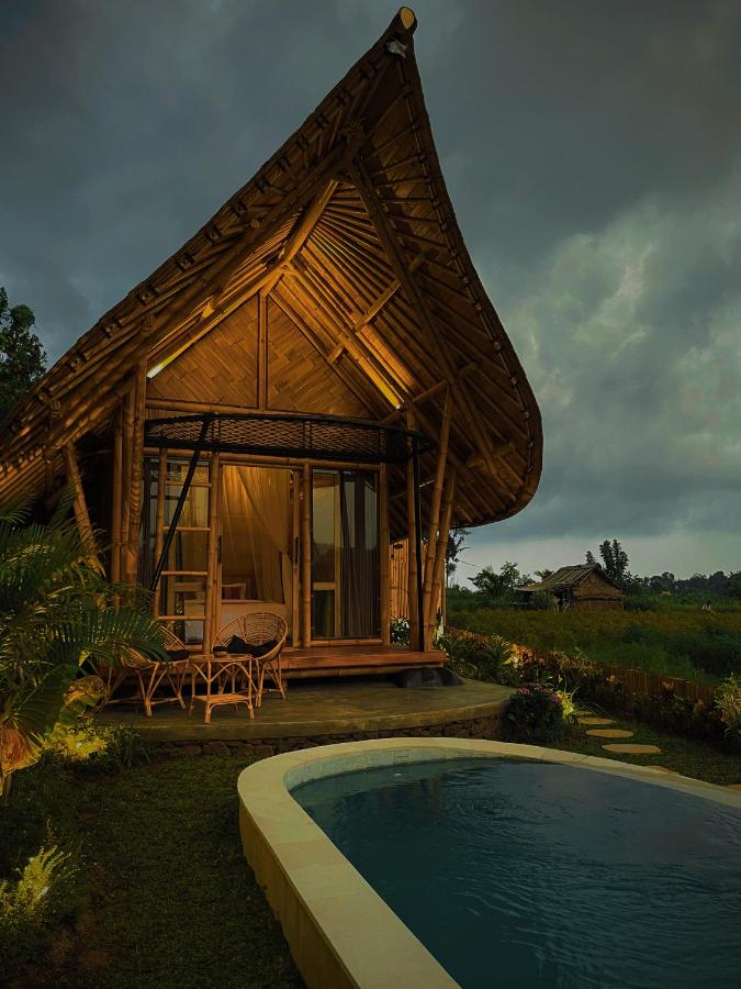 A cozy bamboo house with warm lights glows at dusk—one of the best family villas in Bali—featuring a curved roof, small patio, stone path to a pool, and lush greenery stretching into distant fields under a cloudy sky.