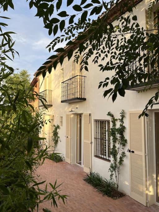 25 Weekend Trips from Madrid Max. 2h Away 59 A sunlit, cream-colored building with shuttered windows and small balconies is partly shaded by leafy branches—typical of charming towns perfect for weekend trips from Madrid. Potted plants and vines grow along the brick walkway in front of the doors.