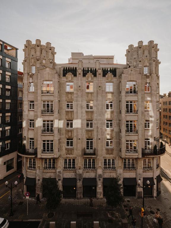 A symmetrical, beige stone building with decorative art deco details, large windows, and small balconies stands on a city corner—much like some boutique hotels in Asturias—surrounded by modern buildings and bustling streets under a clear sky.