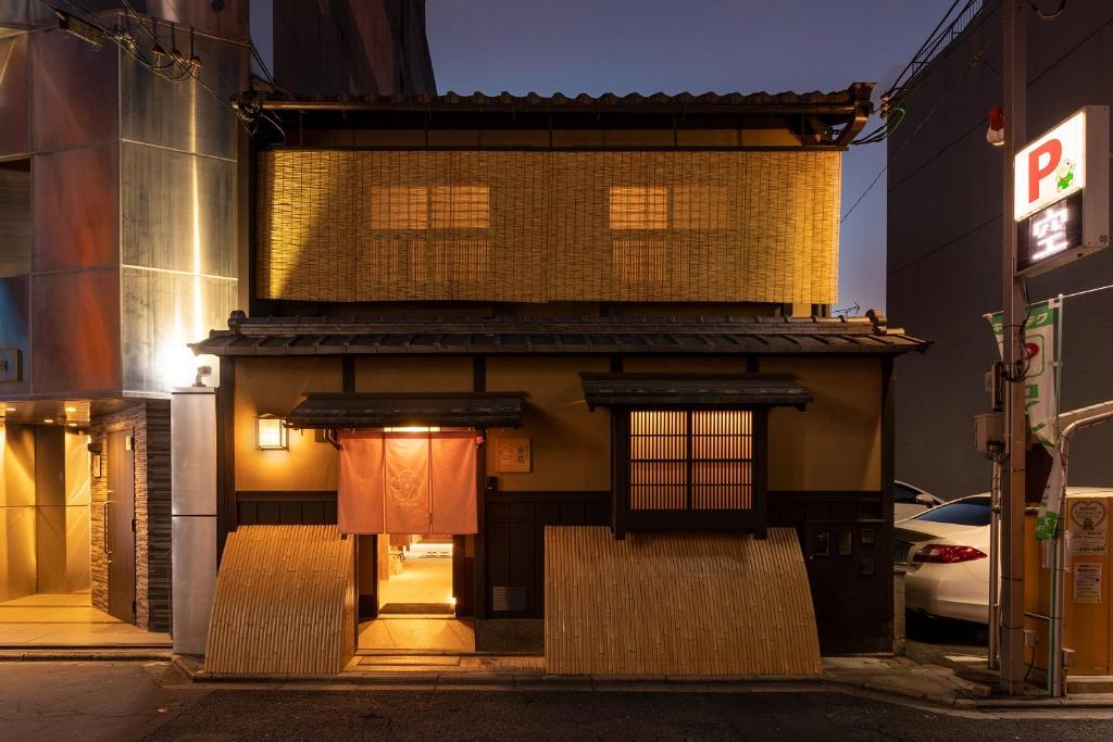 7 UNIQUE Ryokans in Gion Kyoto 27 A traditional Japanese building with bamboo blinds and wooden accents, warmly lit at dusk. A pink noren curtain hangs at the entrance, typical of ryokans in Gion Kyoto, with a modern parking lot sign visible on the right.