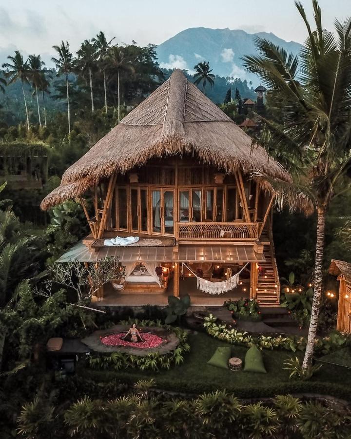 A two-story bamboo house with a thatched roof is surrounded by lush tropical greenery. A woman sits on a flower petal arrangement in the garden—an inviting scene at one of the best family villas in Bali, with palm trees and mountains beyond.