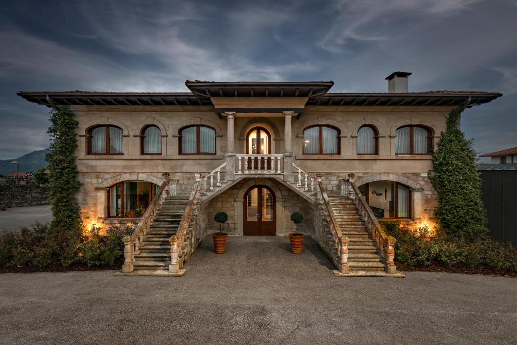 A large stone house with arched windows, twin staircases to a grand entrance, potted plants, and ivy-clad walls evokes the charm of boutique hotels in Asturias, set against dramatic evening lighting and a cloudy sky above.