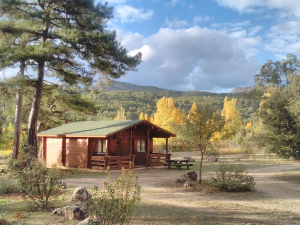 25 Weekend Trips from Madrid Max. 2h Away 82 A small wooden cabin with a green roof sits among trees and bushes, perfect for weekend trips from Madrid. With mountains in the background and a picnic table nearby, the peaceful scene is surrounded by nature.
