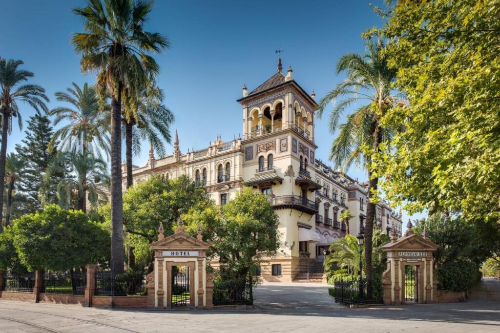 10 UNIQUE Hotels with Pool in Seville 25 A grand historic hotel with a tall central tower, ornate details, and a tiled roof sits among lush palm trees under a clear blue sky, viewed from behind iron gates—an iconic choice for those seeking hotels with pool in Seville.