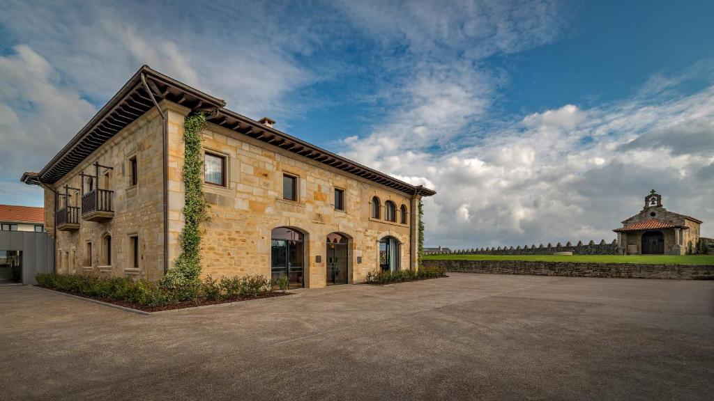 A large stone building with arched windows and a tiled roof stands beside an open paved area, reminiscent of the charm found at boutique hotels in Asturias, with a small chapel and green lawn in the background under a partly cloudy sky.