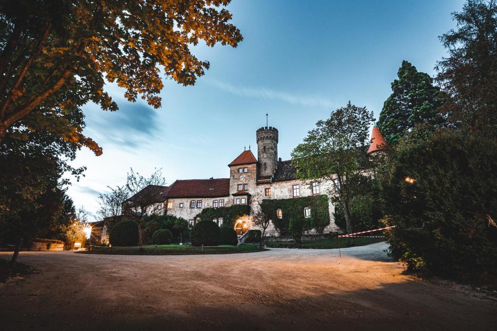 FAIRYTALE Castle Hotels in Bavaria 15 A large historic castle hotel in Bavaria with stone walls and a central tower, surrounded by trees and lush gardens, is photographed at dusk under a clear blue sky. Warm lights illuminate some windows and pathways.