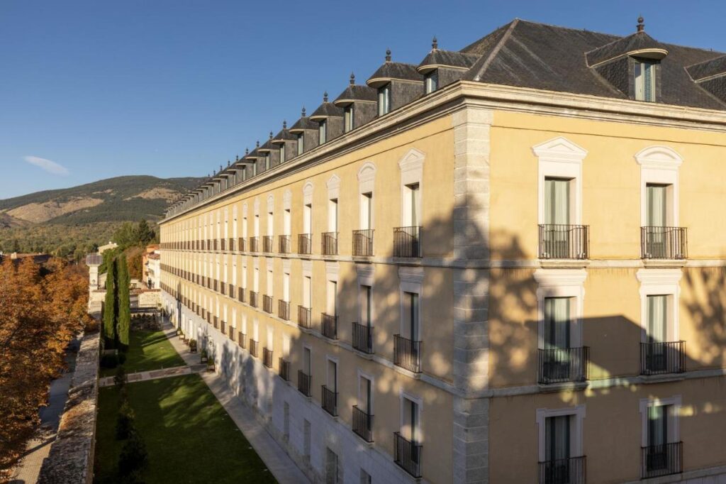25 Weekend Trips from Madrid Max. 2h Away 93 A large, elegant building with yellow walls, black railings, and rows of windows sits beside a green lawn with trees—an ideal spot for weekend trips from Madrid, with hills and blue sky in the background.