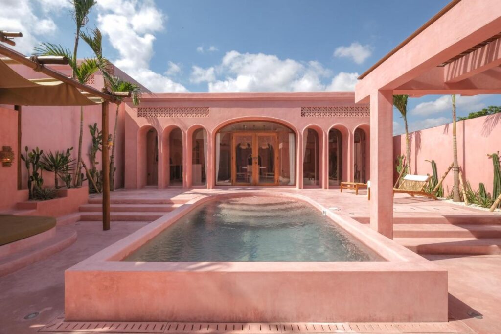 A modern pink villa with arched doorways and windows surrounds a rectangular swimming pool, with palm trees and plants along the walls—one of the best family villas in Bali under a blue sky with scattered clouds.