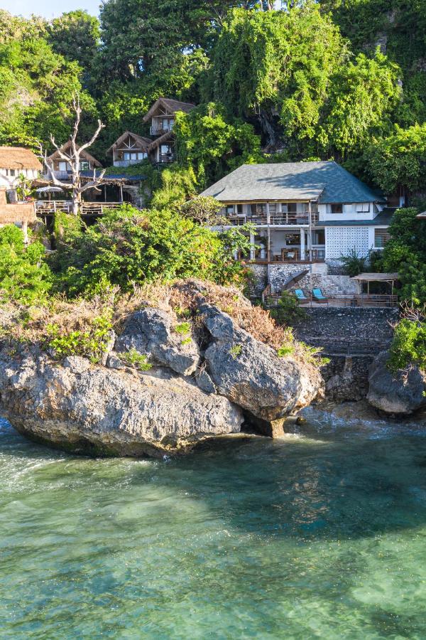 A seaside view of rustic wooden houses, reminiscent of the best family villas in Bali, surrounded by lush green trees and perched on rocky cliffs above clear turquoise water.