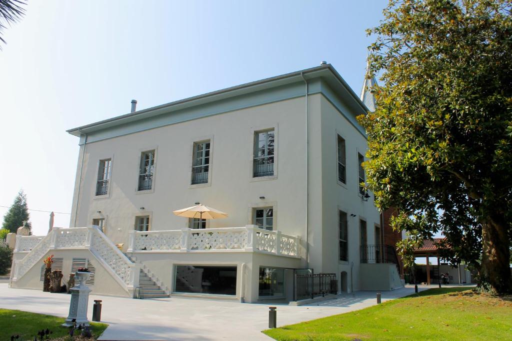 A large, two-story white house with multiple windows and a spacious patio featuring an umbrella and outdoor seating, reminiscent of boutique hotels in Asturias, surrounded by green lawn and trees under a clear blue sky.