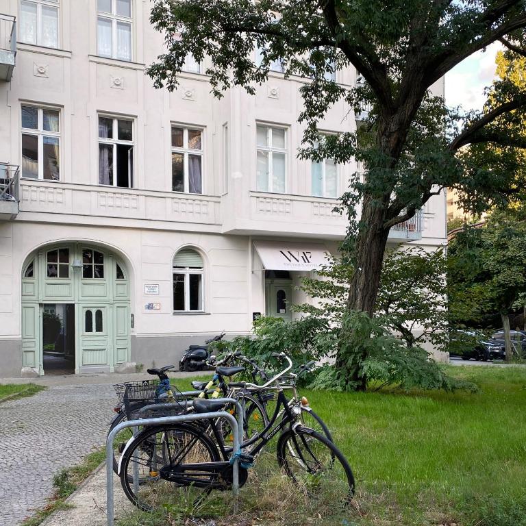 Boutique Hotels in Berlin Kreuzberg 7 Several bicycles are parked at a bike rack on a sidewalk next to a grassy area with a large tree, in front of an apartment building reminiscent of boutique hotels in Berlin Kreuzberg, featuring arched doorways and tall windows.