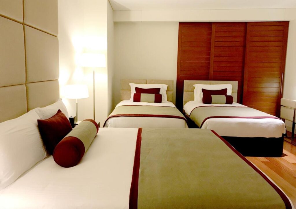 Where to Stay in Doha, Qatar: Transit, City & Beach Hotels 20 A stylish hotel room, perfect for those seeking where to stay in Doha, offers one queen bed and two twin beds with crisp white bedding, brown and beige accents, decorative pillows, and a cozy lamp near wooden sliding doors.