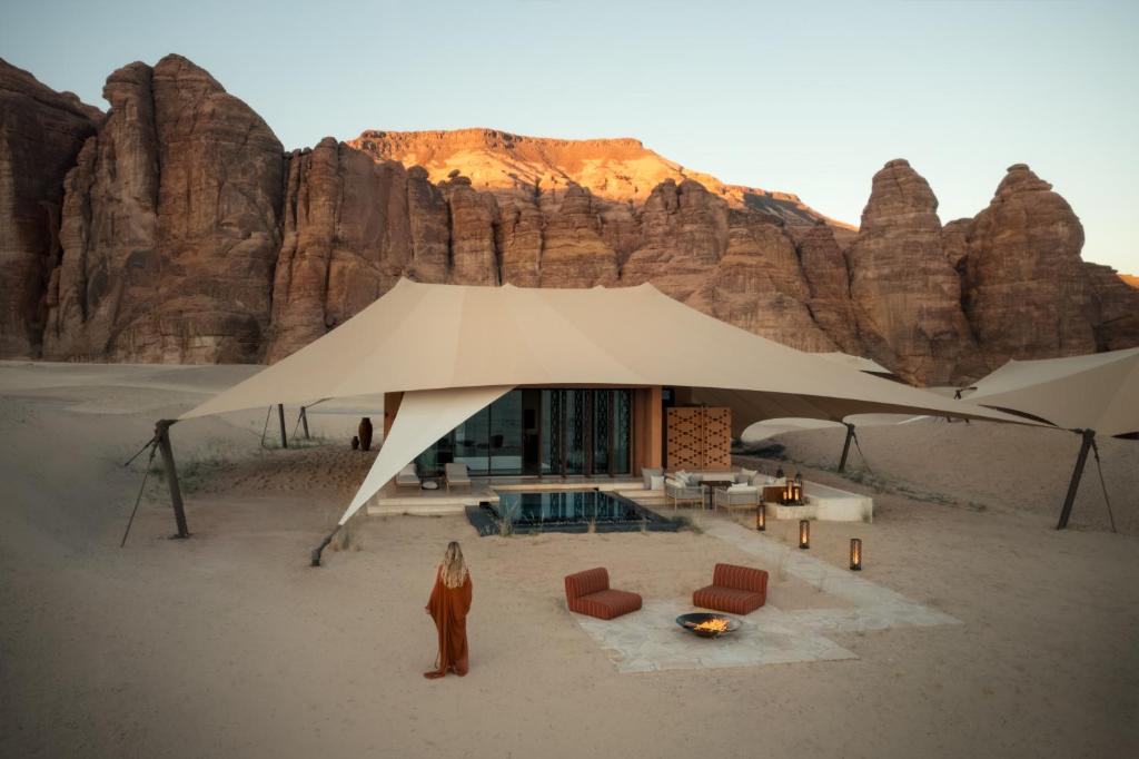 9 Best Hotels in Saudi Arabia: Unique Stays Across the Country 17 A person in a long robe stands near a modern desert tent with outdoor seating, a fire pit, and a pool—an enchanting scene reminiscent of the Best Hotels in Saudi Arabia, set against dramatic rocky cliffs at sunset.