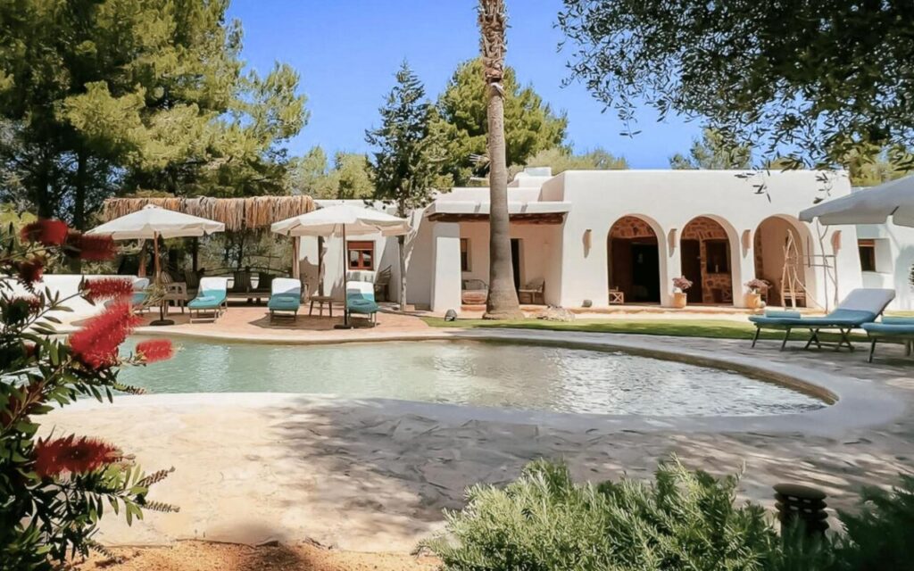 10 AUTHENTIC Finca Hotels in Ibiza 40 A white villa with arched doorways and shaded patios, inspired by finca hotels in Ibiza, is surrounded by trees. In front, a lagoon-shaped pool with lounge chairs and umbrellas sits amid blooming red flowers in the foreground.