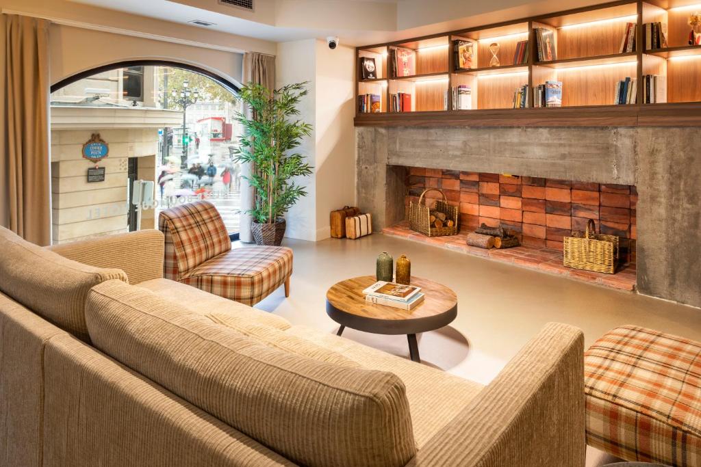 8 Unique Boutique Hotels in Bilbao Spain 12 A cozy living room reminiscent of boutique hotels in Bilbao, with a beige sofa, plaid armchairs, a round wooden coffee table, a large empty brick fireplace, built-in bookshelves, a potted plant, and a large arched window letting in natural light.
