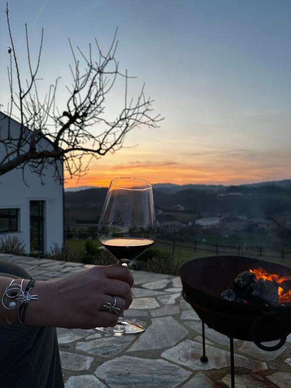 A hand holding a glass of red wine is in the foreground, with a fire pit and scenic hills glowing under a colorful sunset sky—an inviting glimpse into evenings at boutique hotels in Asturias, with a bare tree and part of a building to the left.