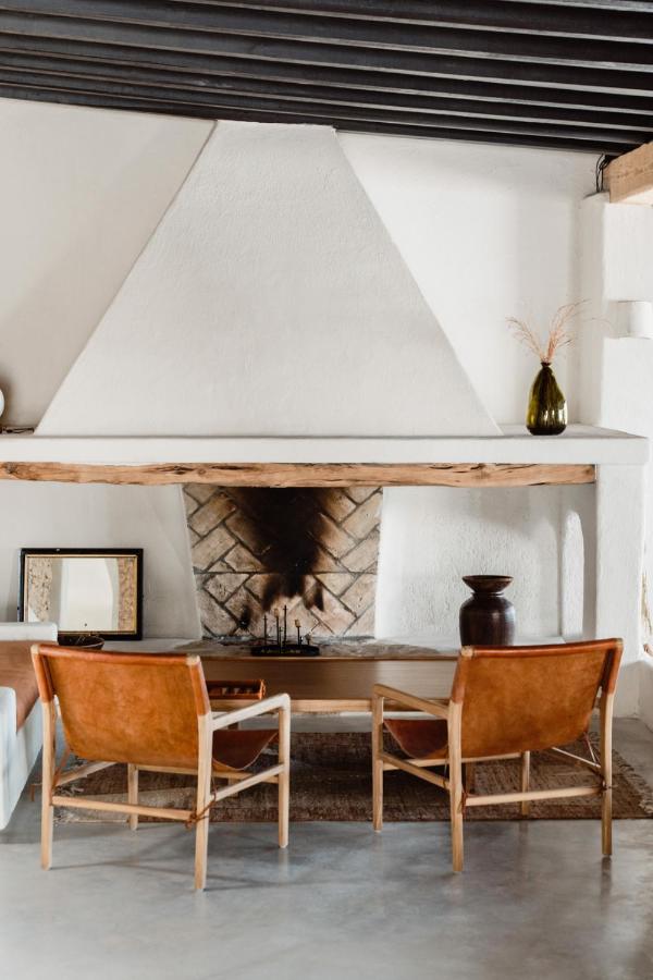 10 AUTHENTIC Finca Hotels in Ibiza 19 A cozy living room features a white fireplace with a wooden mantel, tan leather chairs, and rustic decor reminiscent of finca hotels in Ibiza. Framed art, a dark vase, and dried grass add minimalist charm to the inviting space.