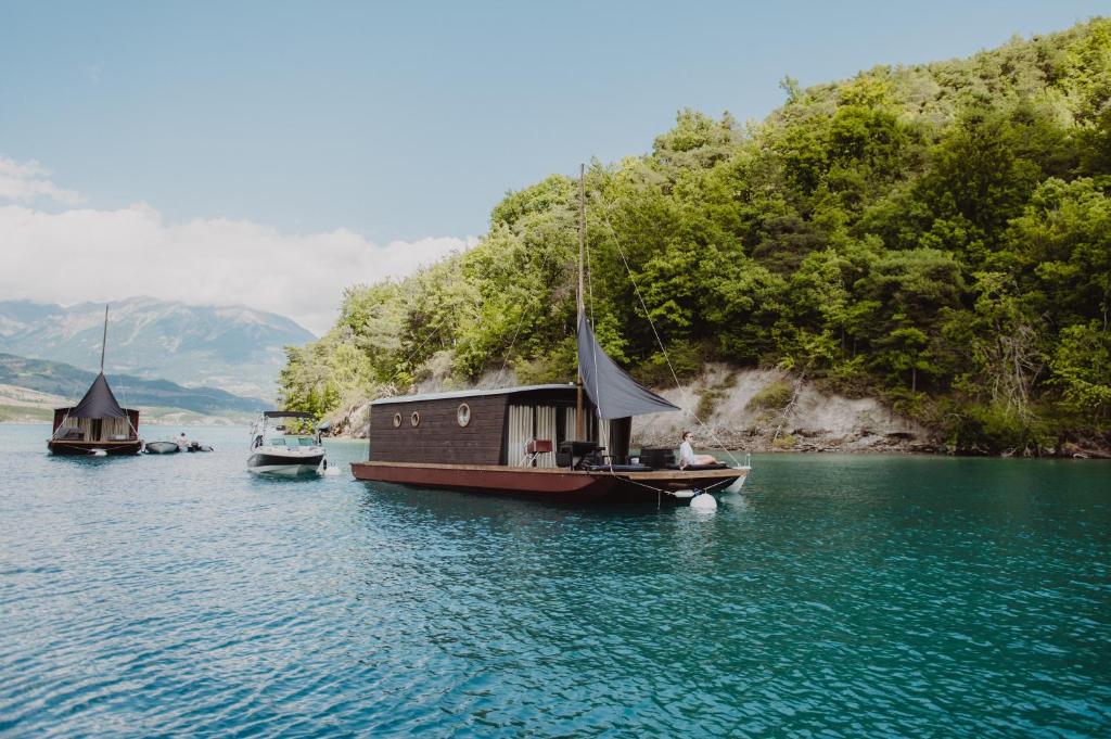 11 Unique Places to Stay in Provence 18 Two houseboats and a motorboat float on a calm blue lake near a forested shoreline, with mountains in the background under a partly cloudy sky—one of the unique places to stay in Provence.