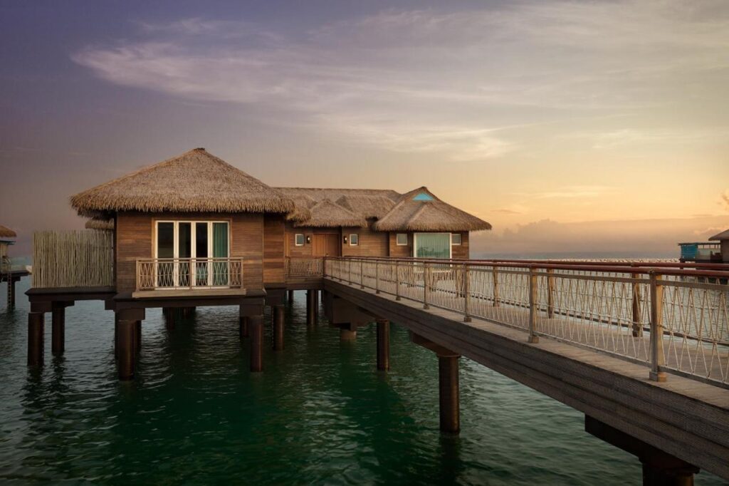 Where to Stay in Doha, Qatar: Transit, City & Beach Hotels 13 A wooden overwater bungalow with a thatched roof stands on stilts above calm green water, connected to land by a walkway—an idyllic escape reminiscent of where to stay in Doha, with a warm sunset sky in the background.