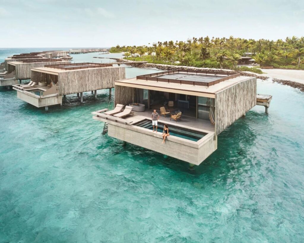 Best Luxury Hotels in Maldives for Your Dream Vacation 1 Luxury overwater villas with private pools sit above clear turquoise water; two people relax on the deck of one villa, surrounded by palm trees and tropical scenery—hallmarks of the best luxury hotels in Maldives—under a partly cloudy sky.