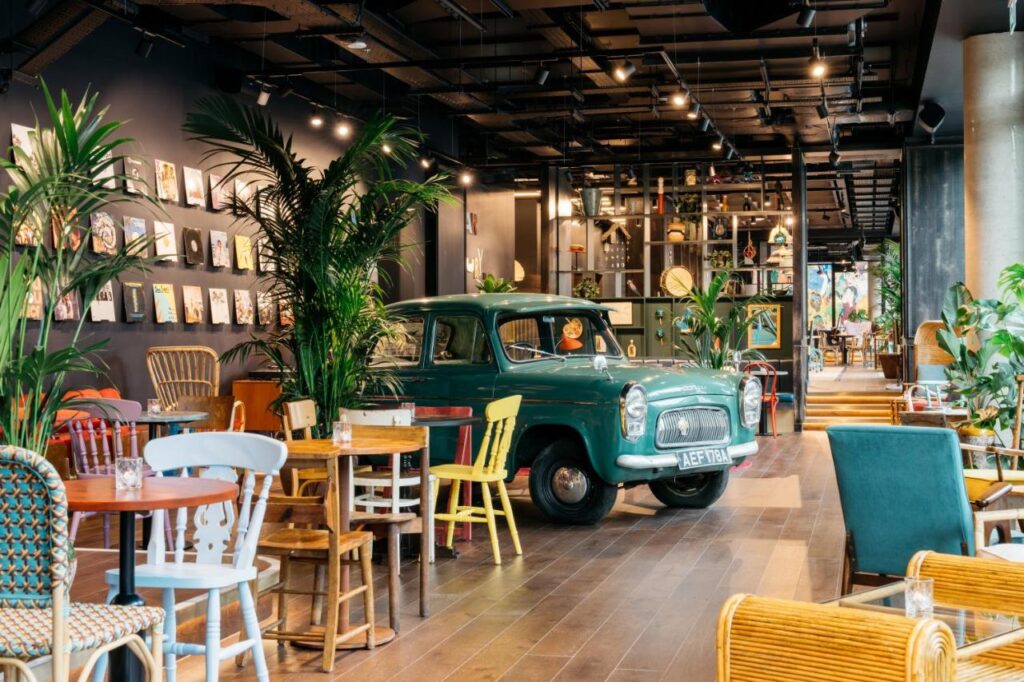 15 Best Boutique Hotels in London, UK 21 A cozy café interior reminiscent of the best boutique hotels London offers, with eclectic furniture, potted plants, and a vintage green car parked inside. Art and books decorate the walls, creating a quirky and inviting atmosphere.