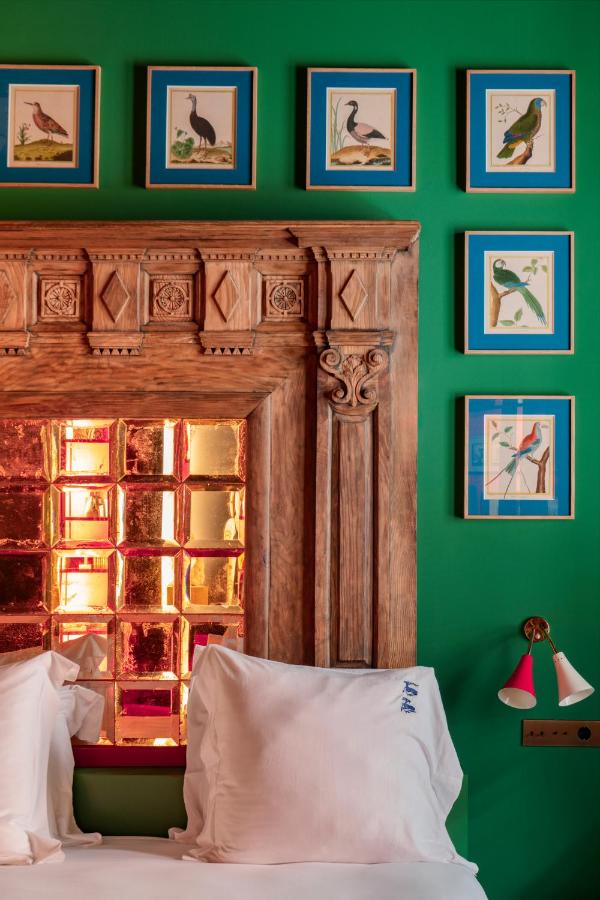 10 UNIQUE Hotels with Pool in Seville 36 A cozy bed with white pillows sits beneath a carved wooden headboard and mirrored panel, set against a green wall decorated with framed bird illustrations—perfect for relaxing after exploring hotels with pool in Seville.