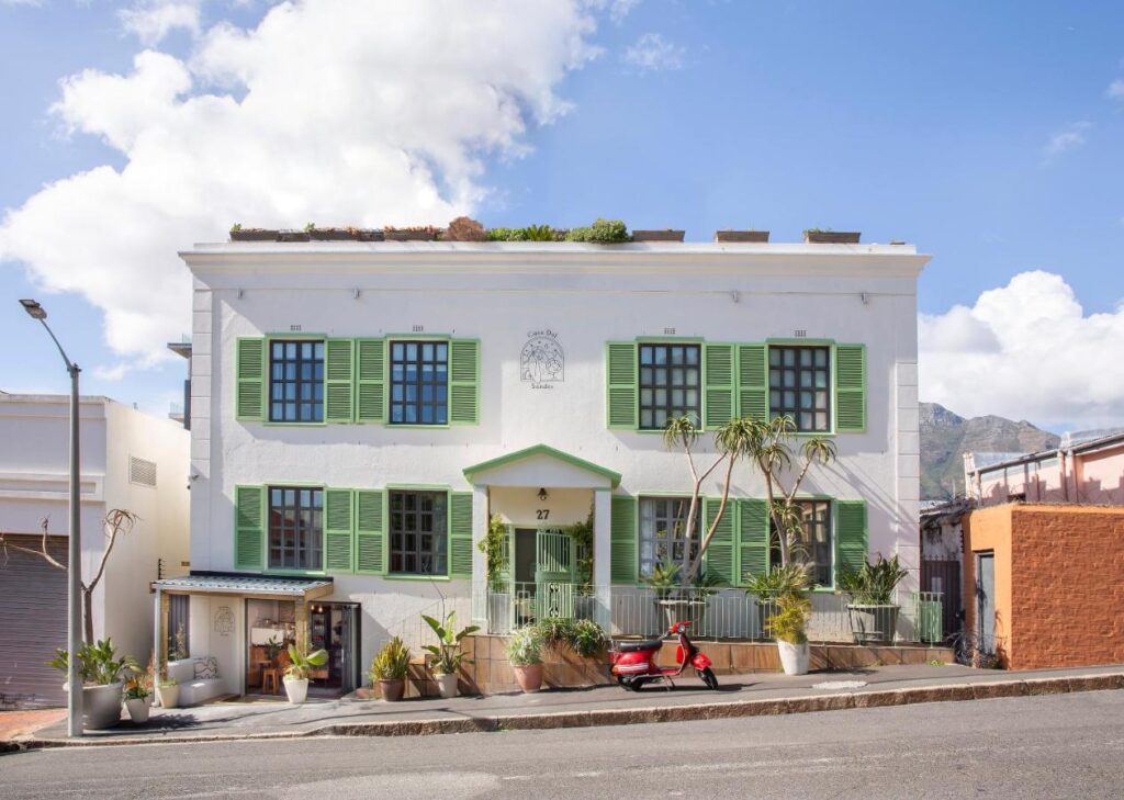 Unique Places to Stay in Cape Town 2 A white two-story building with green shutters and a small porch, surrounded by potted plants and trees, stands out among unique places to stay in Cape Town. A red scooter is parked in front, under a blue sky, on a street that slopes downward.