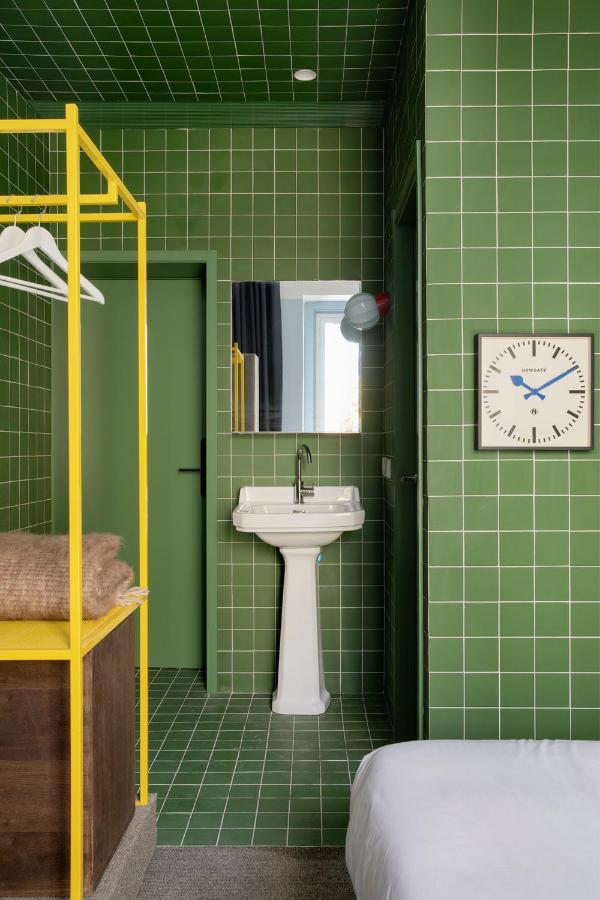 Where to Stay in Madrid: 14 Best Hotels 4 A modern room—perfect for travelers wondering where to stay in Madrid first time—with green tiled walls and floor, a white pedestal sink, a wall clock, mirror, and a yellow open wardrobe with hangers and blankets beside a partially visible bed.