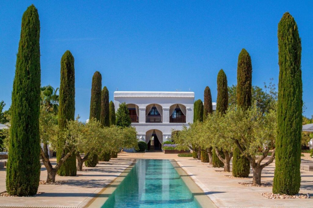 10 AUTHENTIC Finca Hotels in Ibiza 34 A symmetrical courtyard at one of the elegant finca hotels in Ibiza features a long rectangular pool lined with tall cypress and olive trees, leading to a white two-story building with arches and balconies under a clear blue sky.