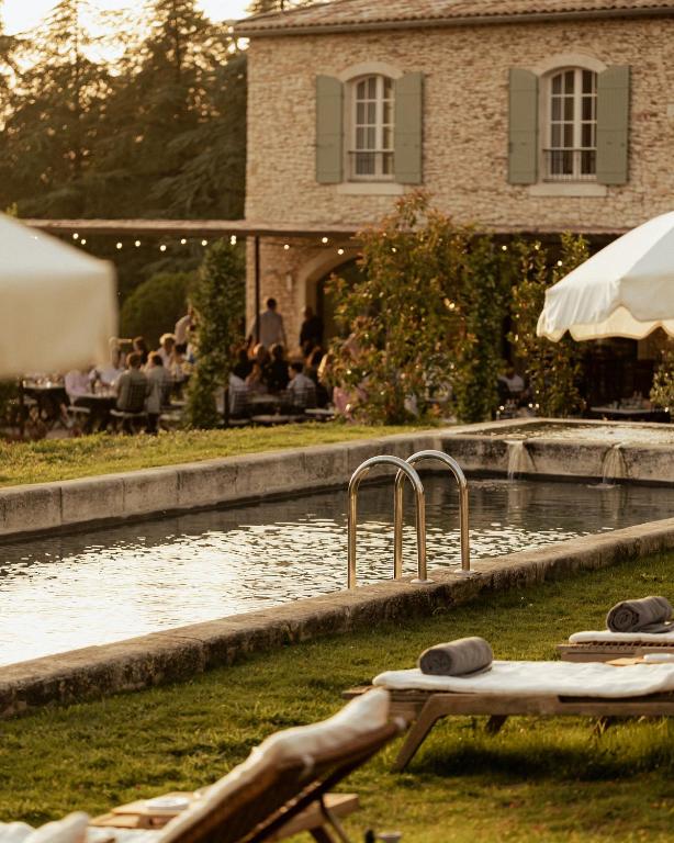 11 Unique Places to Stay in Provence 41 A tranquil outdoor pool with metal steps is bordered by grass and lounge chairs with rolled towels, set in front of a rustic stone building—one of the unique places to stay in Provence—with people dining under string lights.