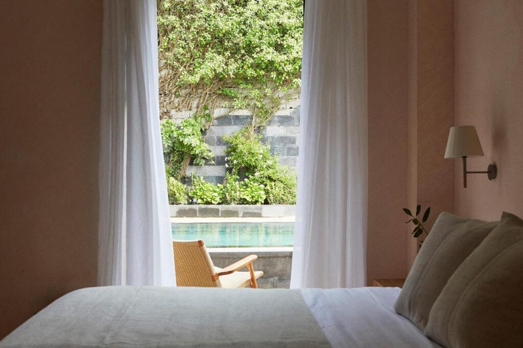 Best Boutique Hotels in San Sebastian 21 A cozy bedroom with white bedding and a light wooden chair beside a window with sheer white curtains, reminiscent of boutique hotels in San Sebastian, overlooking a pool and lush greenery outside.