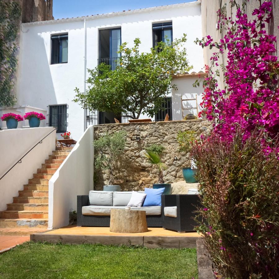 A cozy outdoor patio, reminiscent of boutique hotels in Costa Brava, features a sofa, cushions, and a tree stump table nestled in a garden with stone steps, white building backdrop, pink flowers, and lush green grass.