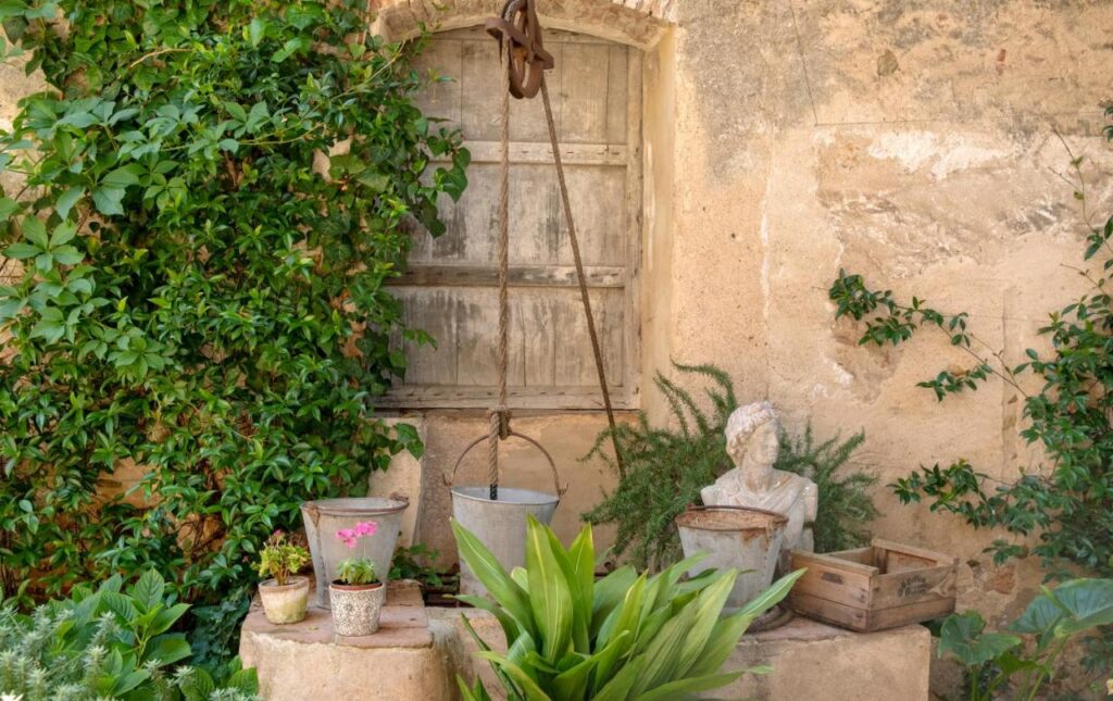 A rustic stone wall with a closed wooden window, a pulley holding a metal bucket, potted plants, and green foliage creates a quaint, serene garden scene reminiscent of boutique hotels in Costa Brava.