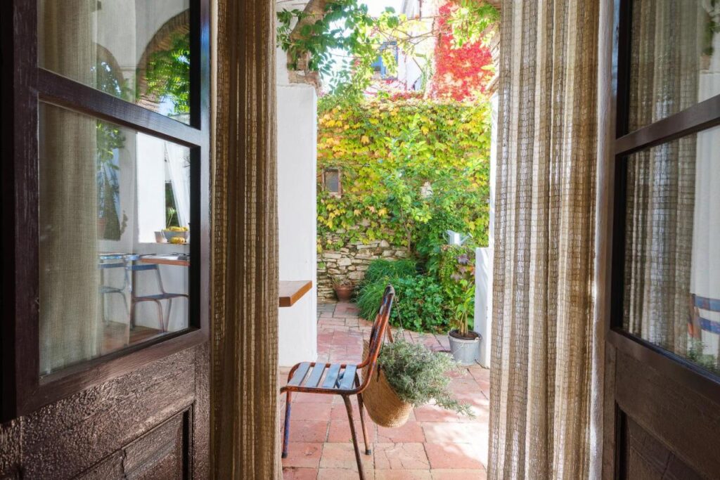 View through sheer curtains and an open doorway onto a sunlit patio with a rustic metal chair, potted plants, and colorful ivy-covered walls in a cozy, Mediterranean-style courtyard reminiscent of boutique hotels in Costa Brava.