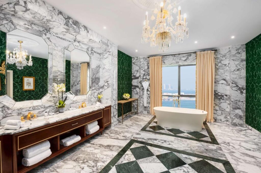 Where to Stay in Doha, Qatar: Transit, City & Beach Hotels 31 Luxurious bathroom with marble floors and walls, large mirrors, a double vanity, and standalone bathtub by a window with ocean view—perfect inspiration if you're searching for where to stay in Doha. Green wallpaper accents and chandeliers enhance the elegance.