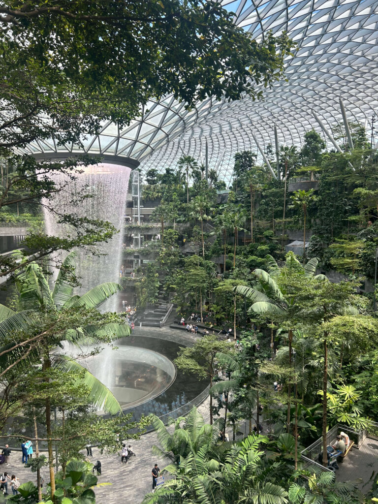 13 Most Unique Hotels in Singapore for an Incredible Holiday 2 A large indoor garden with lush greenery, a tall waterfall cascading from the ceiling into a circular pool, and people walking on pathways under a glass dome roof—one of the highlights of unique hotels in Singapore.