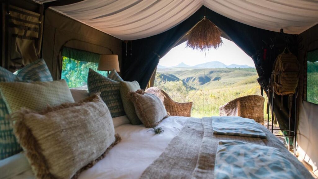 View from inside a cozy glamping tent with a bed, cushions, and wicker chairs facing the open entrance—perfect for enjoying grassy hills and distant mountains under a bright sky, reminiscent of Game Lodges in Western Cape.