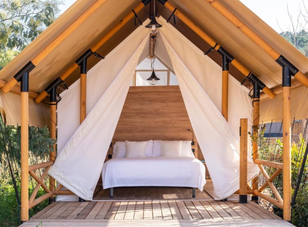 A cozy glamping tent with open canvas flaps reveals a neatly made bed with white linens. Reminiscent of boutique hotels in Costa Brava, the wooden frame and deck blend seamlessly into the natural outdoor environment.