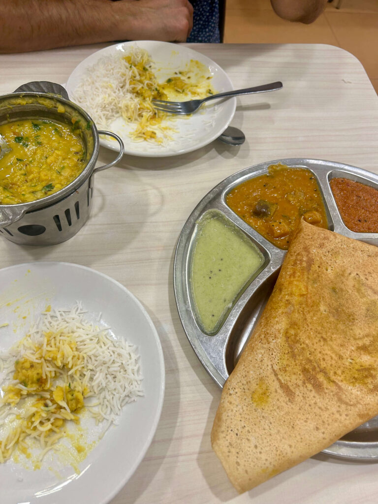 13 Most Unique Hotels in Singapore for an Incredible Holiday 61 Two plates on a table: one with rice and yellow lentil curry, the other with a dosa and three chutneys—reminiscent of breakfasts served at unique hotels in Singapore. A hand and arm are visible, with a small pot of curry beside the plates.