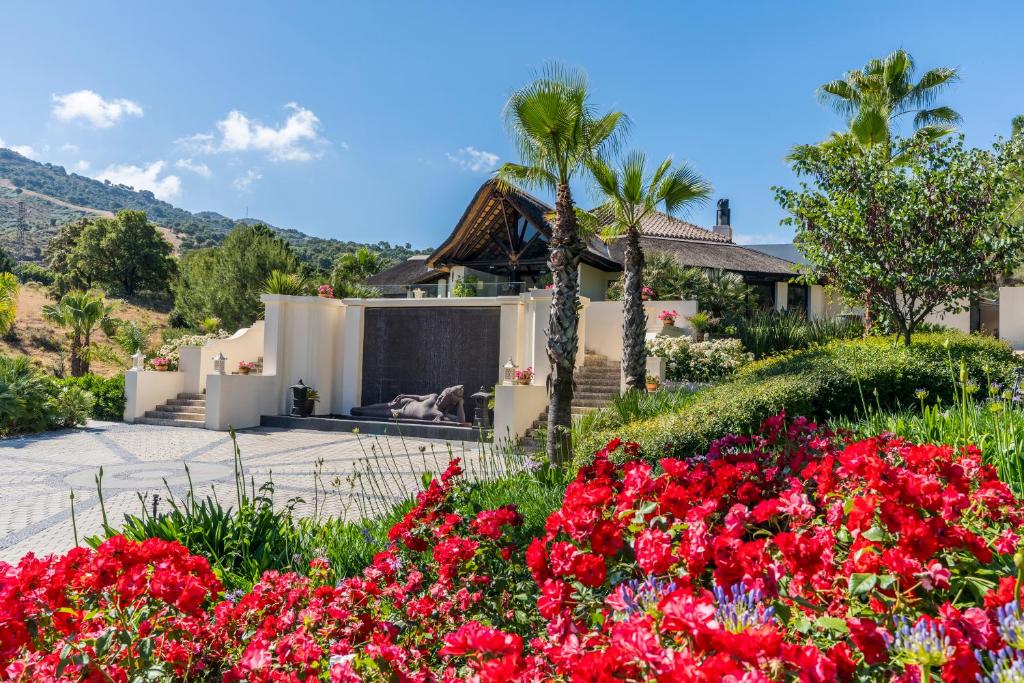 Adults-Only Hotels in Costa del Sol 27 A luxurious house with a tile roof is surrounded by lush greenery and vibrant red flowers, reminiscent of the serene ambiance found at adult only hotels in Costa del Sol, all set against rolling hills beneath a bright blue sky.