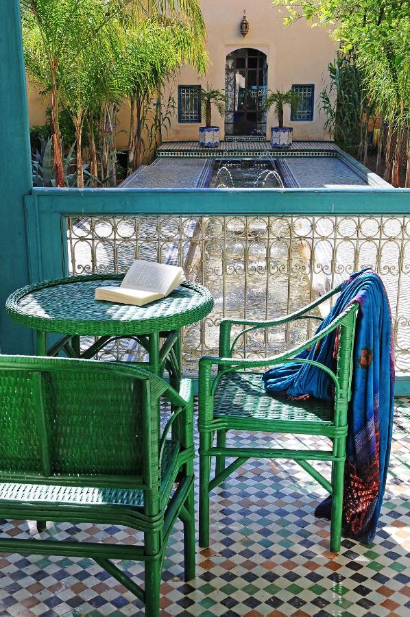 7 UNIQUE Riads in Fez You Won't Forget 13 A sunlit patio with green wicker chairs and a table, holding an open book and a colorful scarf. Beyond the railing, like many riads in Fez, a courtyard features a long fountain, palm trees, and a tan building with blue-trimmed windows.