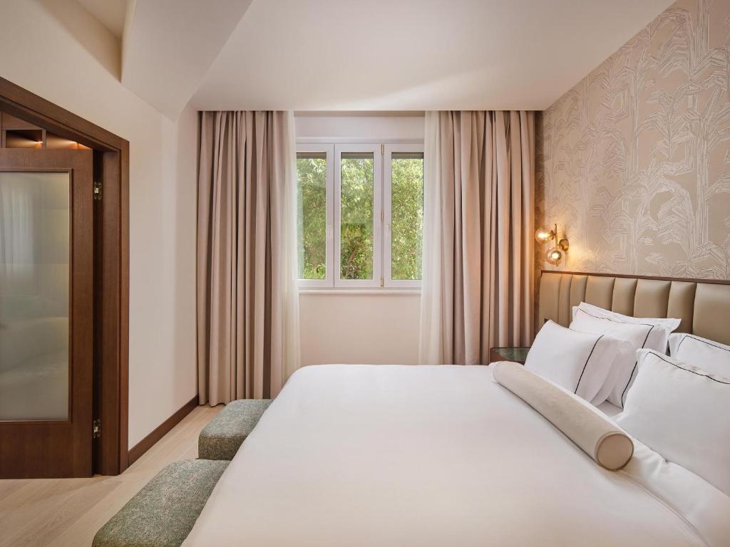 8 TOP Boutique Hotels in Split 31 A bright, modern hotel room in the style of boutique hotels in Split, featuring a large bed with white bedding, decorative pillows, two small benches at the foot, light-colored curtains, patterned wallpaper, and a window overlooking greenery.