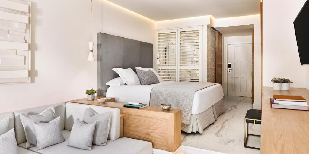 Adults-Only Hotels in Costa del Sol 12 A modern hotel room with a large bed, gray upholstered headboard, white bedding, a small couch with gray pillows, wooden accents, and minimal decor—ideal for those seeking adult only hotels in Costa del Sol. A TV is mounted on the wall opposite the bed.