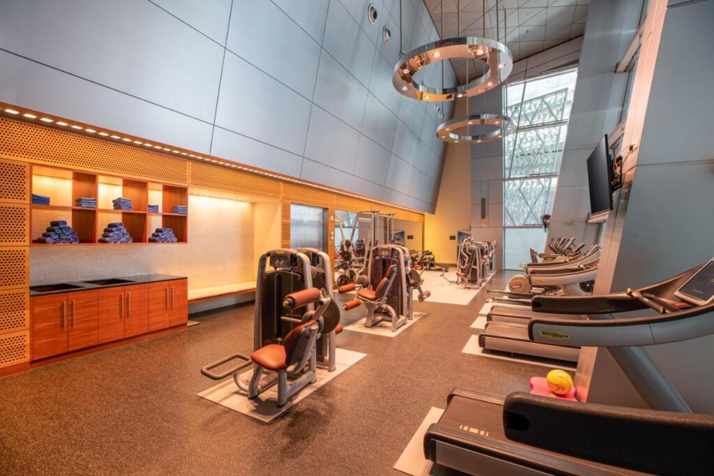 Where to Stay in Doha, Qatar: Transit, City & Beach Hotels 22 Modern gym with cardio machines, weight machines, and a row of treadmills facing a large window—an excellent choice for those wondering where to stay in Doha. Shelves with folded towels line the left wall, and circular ceiling lights hang overhead.