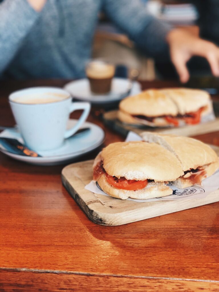 1 day in Seville Spain: My Favorite City 12 Two sandwiches with tomato and cured meat on wooden boards sit on a wooden table next to a cup of coffee—a classic meal to enjoy during 1 day in Seville, Spain, with someone in a blue shirt reaching for food in the background.