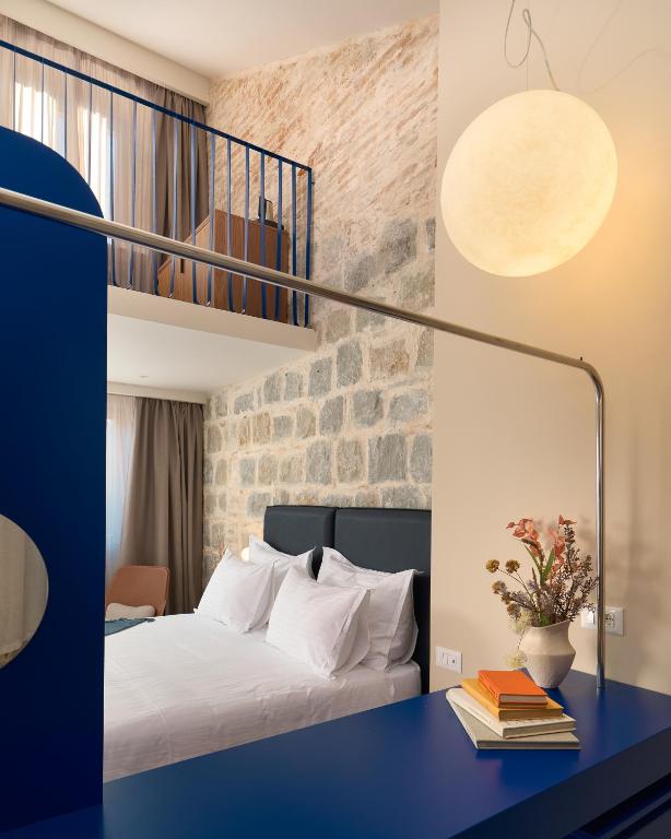 8 TOP Boutique Hotels in Split 17 A modern hotel room—ideal for those seeking Where to Stay in Split—features a loft, stone wall, neatly made bed, blue desk with books and flowers, a large round light fixture, and a window with beige curtains letting in natural light.